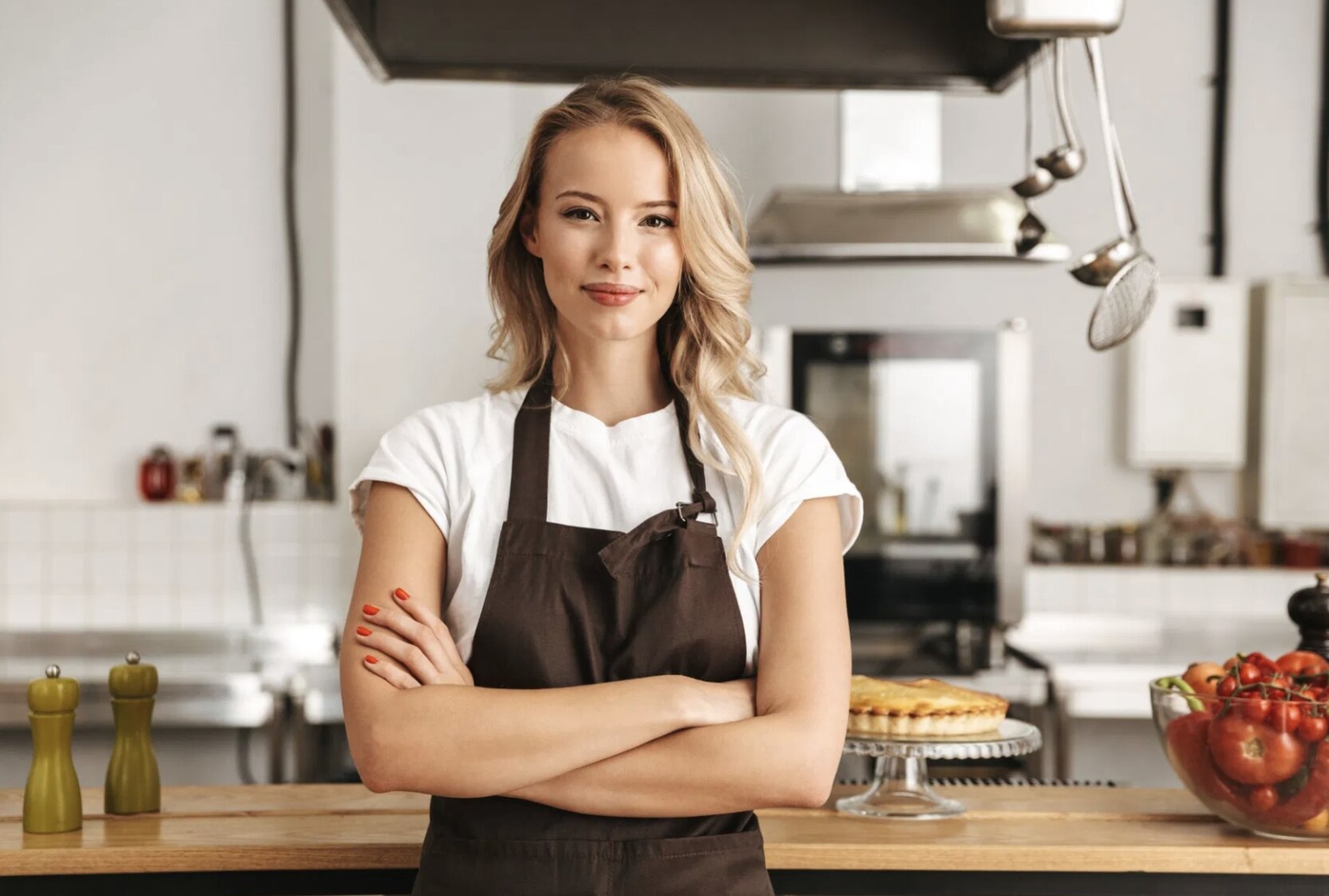 A confident female chef in a brown apron standing in a modern kitchen with her arms crossed, ready for action.